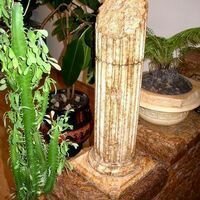 сколотая колонна из камня украшает пьедестал из дикого камня скалистый и красиво смотрится на фоне домашних цветов